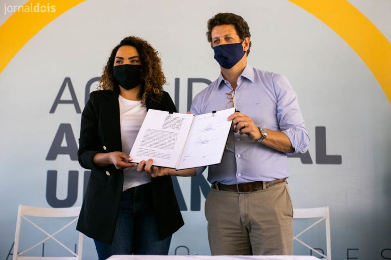 Fotografia mostrando uma mulher negra e um homem branco lado a lado. Ambos seguram um documento aberto e estão fotografados da cintura para cima. A mulher está vestindo um blazer preto, uma blusa branca e uma calça escura. Seus cabelos são cacheados da cor castanho claro. Ela também está de máscara preta. O homem veste calça social bege, camisa social lilás de mangas compridas e uma máscara azul marinho. Em seu colarinho há um óculos de armação vermelha pendurado. Seus cabelos são curtos e da cor castanho escuro.