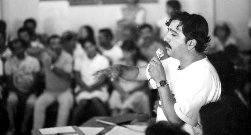 Fotografia em preto e branco que mostra em primeiro plano Chico Mendes, um homem branco de cabelos e bigodes pretos, da cintura para cima, vestindo uma camiseta branca e segurando um microfone enquanto está com o braço direito levantado gesticulando. Em segundo plano há pessoas asssistindo seu discurso.