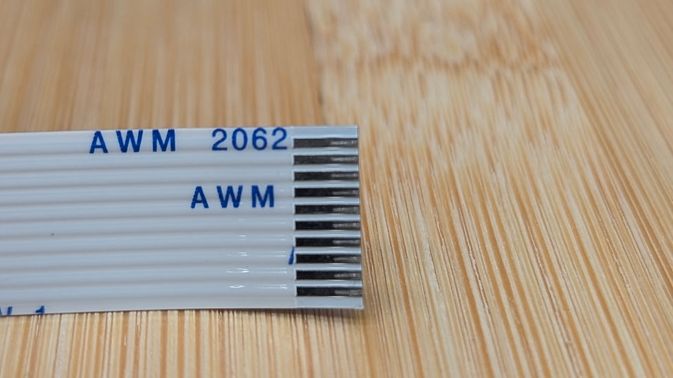Thumbnail: Close-up of the end of a white flat flexible cable (FFC) labeled AWM 2062, showing exposed metal contacts against a light woo