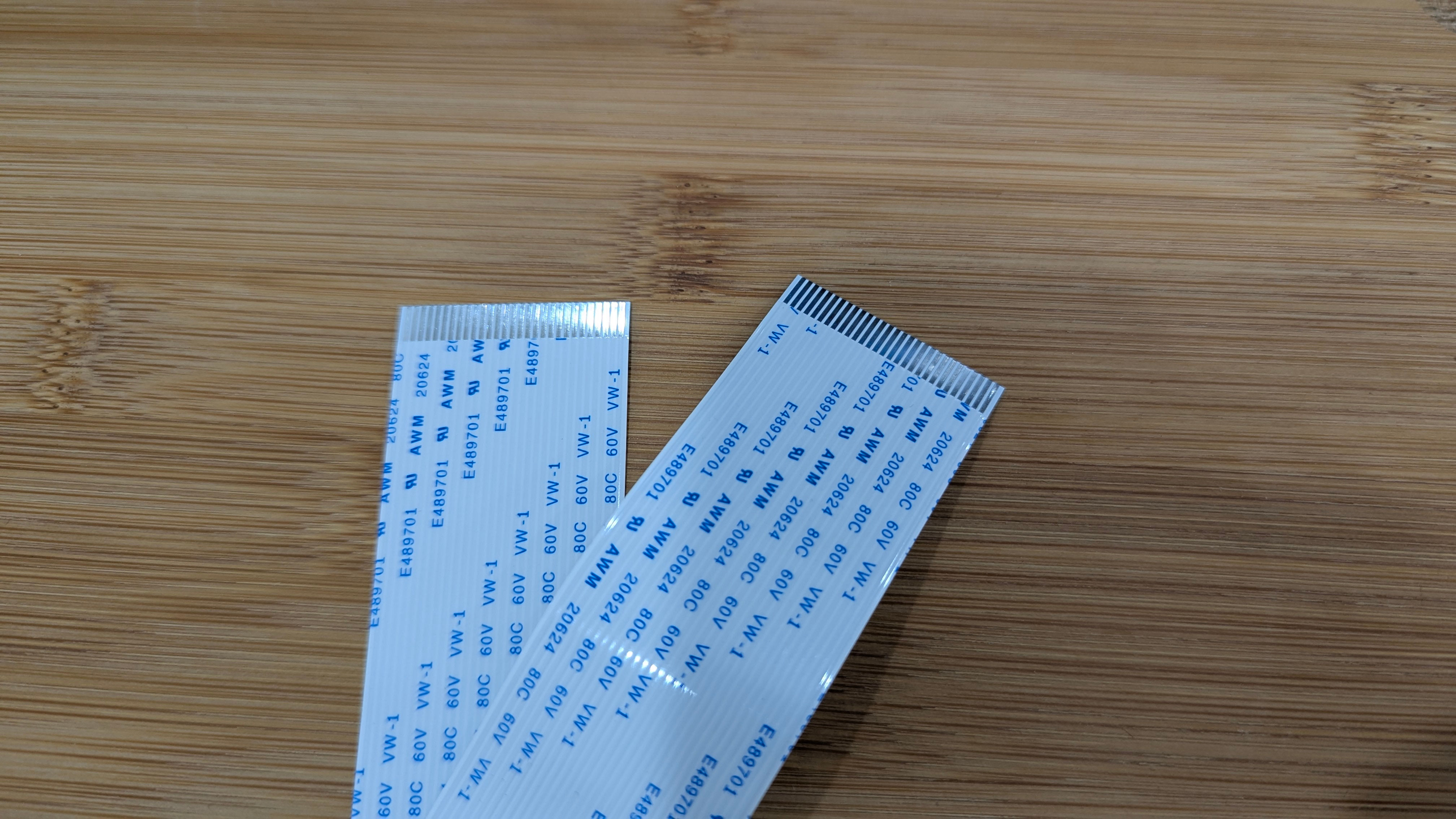 Two flat white ribbon cables with blue text and exposed metal connectors at the ends, lying on a light wooden surface
