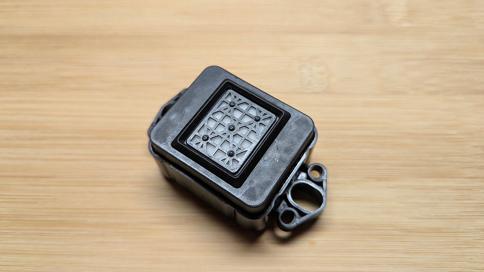 Thumbnail: A small, rectangular black plastic electronic component with a patterned mesh on top sits on a light wooden surface_