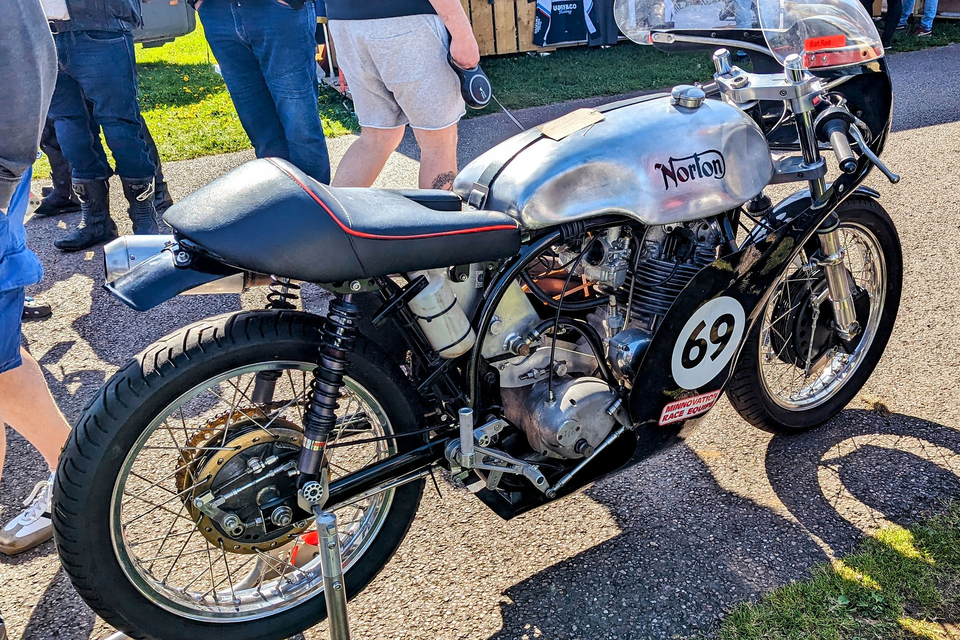 Vintage Norton motorcycle with the number 69 on a sunny day. People in casual attire and boots stand nearby on a paved road.