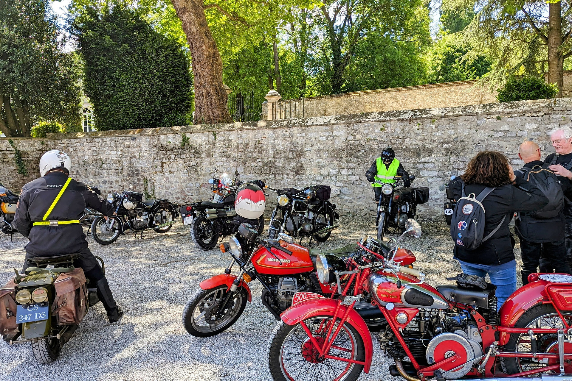 People gather around vintage motorcycles in a sunny, tree-lined parking area. One person wears a hi-vis vest. A stone wall forms the backdrop.