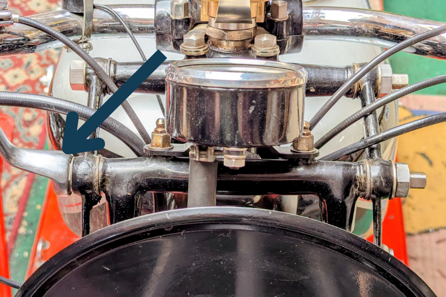 Close-up of a vintage Norton motorcycle handlebar, gauges and girder forks with cables, chrome parts, and a blue arrow pointing to the left fork link. 