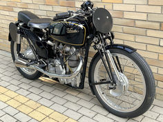 Vintage 1948 black Velocette KTT motorcycle with "Velocette" logo, parked by a tan brick wall on a tiled walkway. Chrome details enhance the classic look.