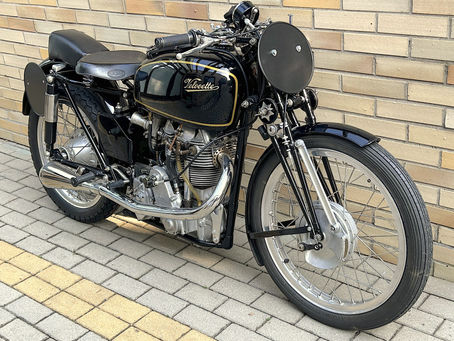 Vintage 1948 black Velocette KTT motorcycle with "Velocette" logo, parked by a tan brick wall on a tiled walkway. Chrome details enhance the classic look.