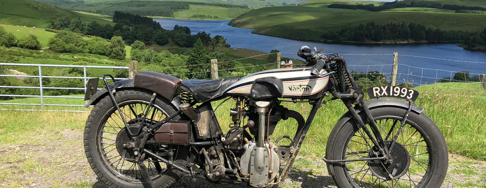 Vintage Norton CS1 motorcycle parked on a hill overlooking a lush valley with a lake. Blue sky, fluffy clouds, and green hills in the background.