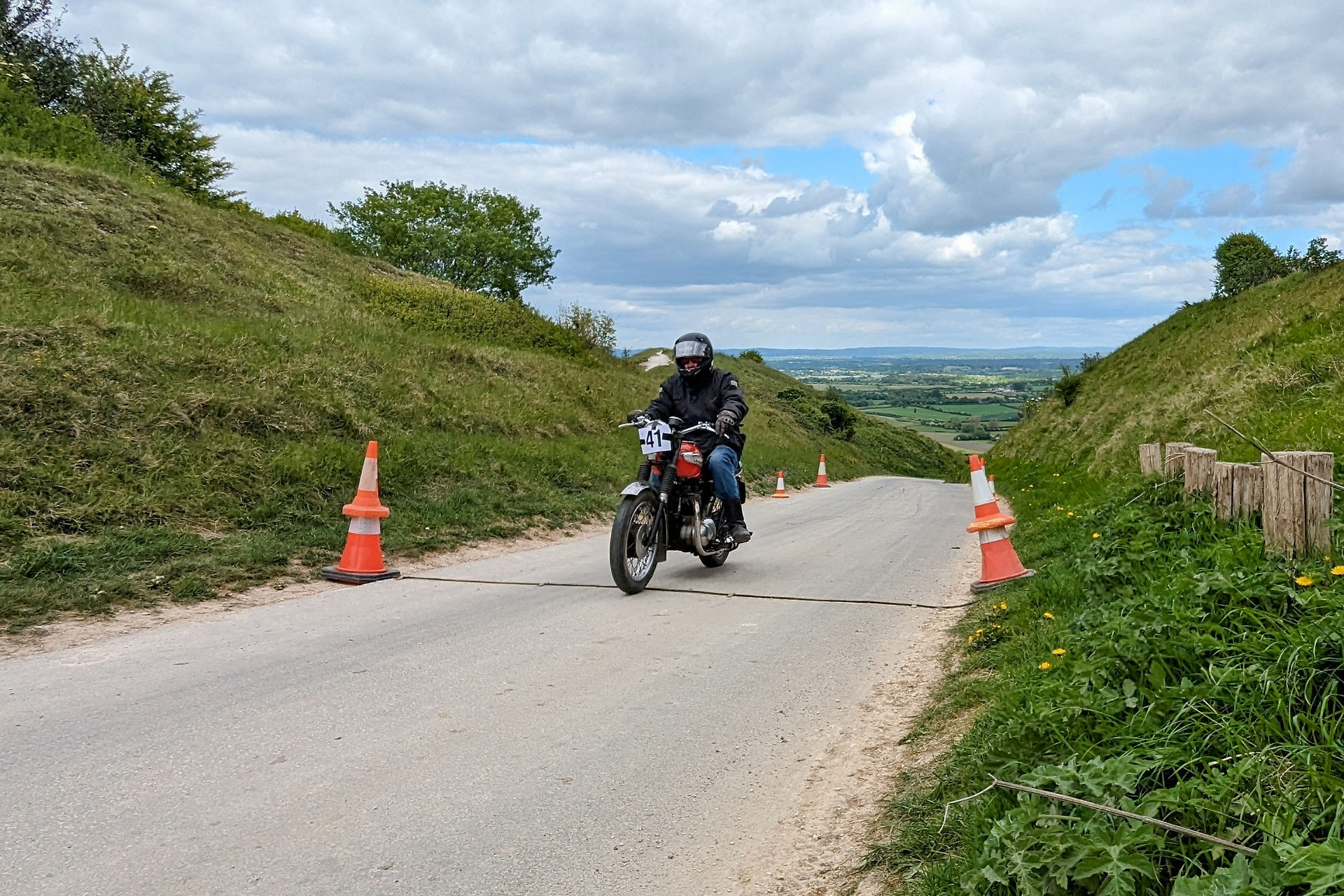 Motorcyclist in helmet rides uphill on a rural road. Orange cones line the path. Greenery and cloudy sky in the background. Number 41 on bike.