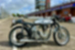 Vintage Norton International motorcycle parked on an empty track at Isle of Man, with cloudy sky and race banners. Red motorcycle in background.