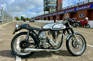 Classic Norton motorcycle parked on an empty Isle of Man race track. Overcast sky, red bike in background, flags and signage visible.