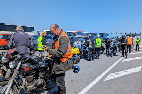 Motorcyclists in high-vis vests gather in a parking lot with buses. A man checks his phone near vintage bikes, under a clear blue sky.