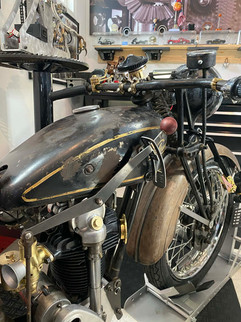 Vintage Velocette motorcycle with a black and gold patina fuel tank, chrome handlebars, and brown leather seat. Checkered floor and framed picture in the background.
