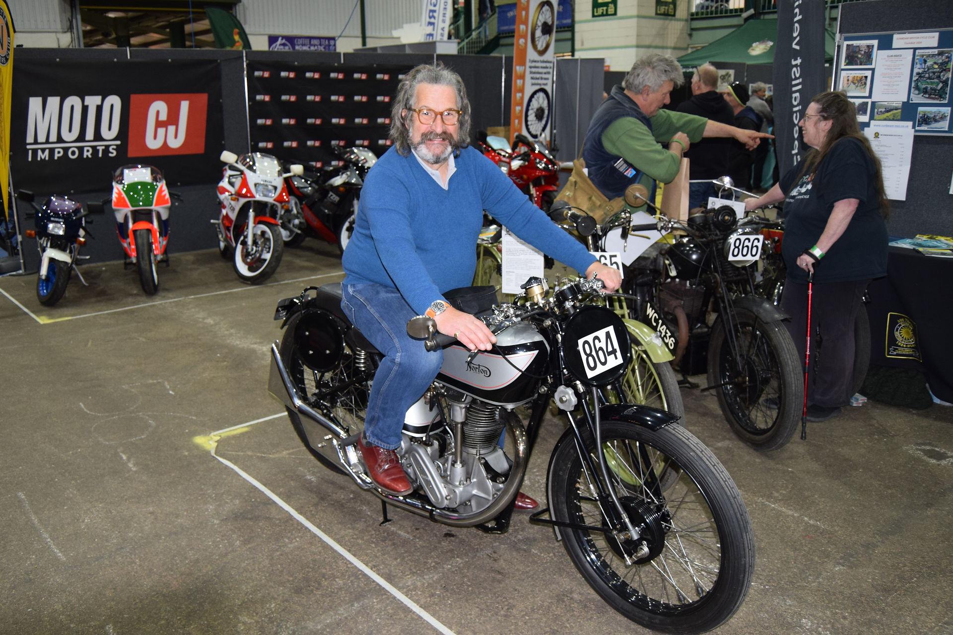 Man in blue sweater sits on a vintage Norton motorcycle with number 864 indoors. Other motorcycles and people in the background.