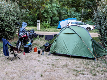 Motorcycle camping setup with green tent, hanging laundry, folding chair, and car in a wooded campsite.