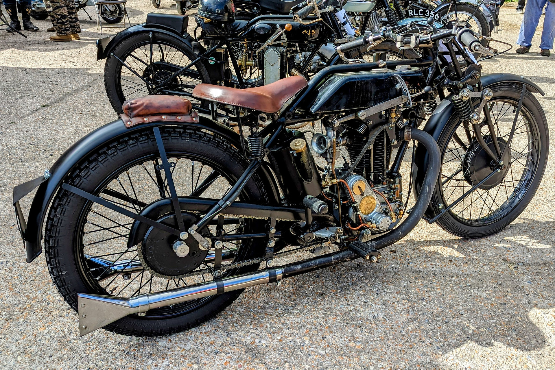 Vintage motorcycle with brown seat displayed on concrete, surrounded by other classic bikes; sunny day, casual gathering.