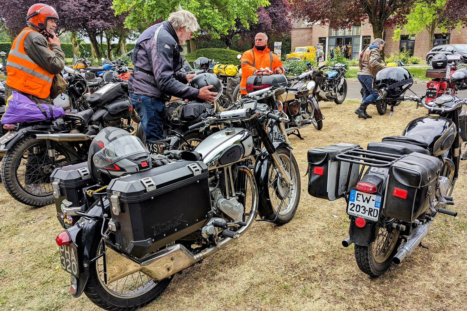 People with vintage motorcycles, two wearing orange jackets. Helmets and bags on bikes. Park setting with trees, grass, and buildings.