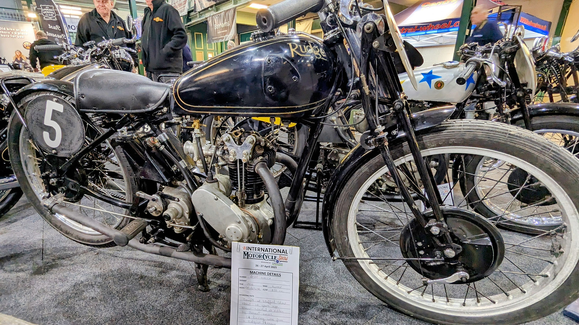 Vintage Rudge motorcycle with a "5" plate displayed at a show. Two people stand behind it. "International Motor Cycle Show" sign in front.