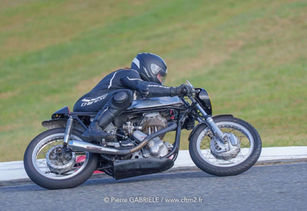 Motorcycle racer in black gear leans into a turn on a track. The bike is silver and chrome with visible engine parts, green grass in background.