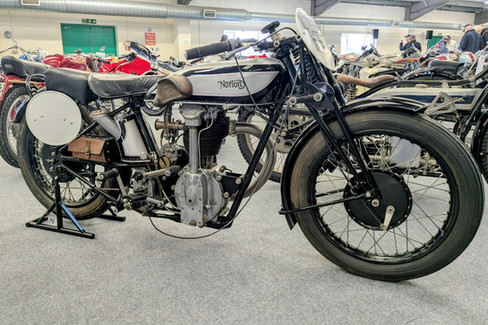 Vintage Norton CS1 motorcycle with silver and black details on display. Surrounded by other classic bikes in an indoor exhibition space.