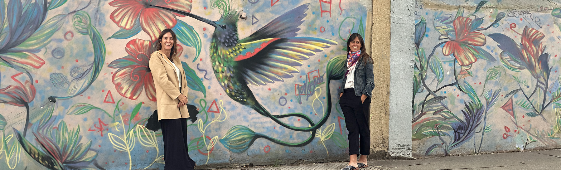 Loli Delger and Majo Garrido, the visionaries behind The Hummingbird Trip, posing before a vibrant hummingbird mural—weaving authentic journeys through the soul of South America.