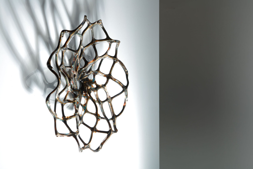 A glass hypotrochoid design sculpture on a wall, colours are earthy tones, background is white.