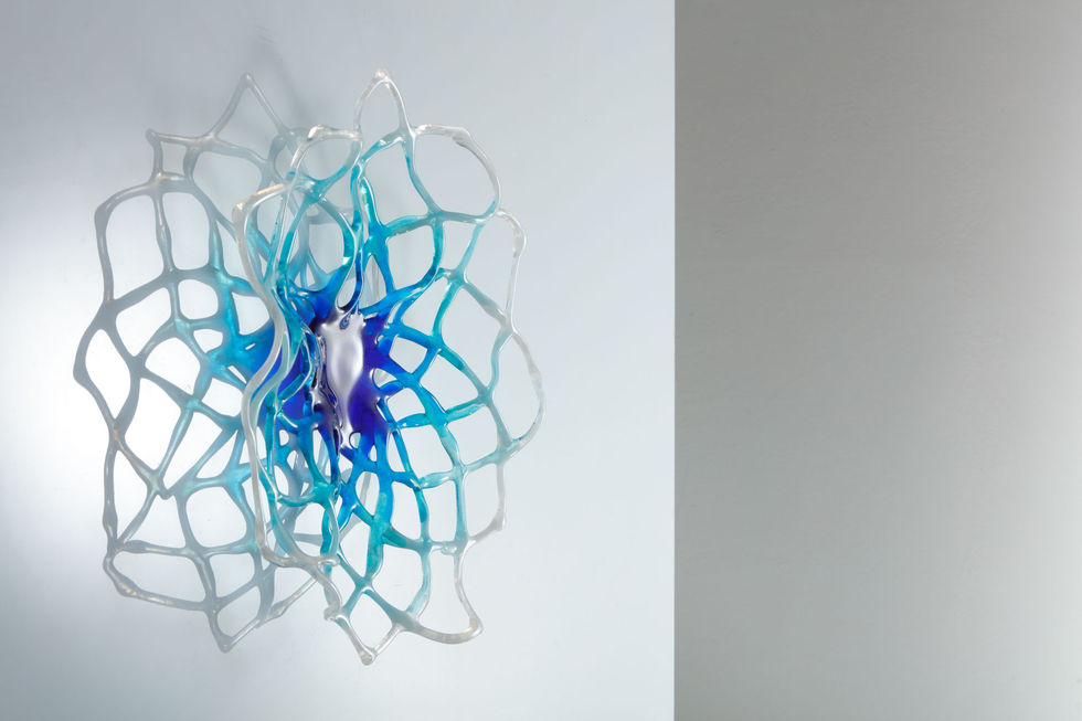 A glass hypotrochoid design sculpture on a wall, colours are blues, aquamarines and white, background is white.
