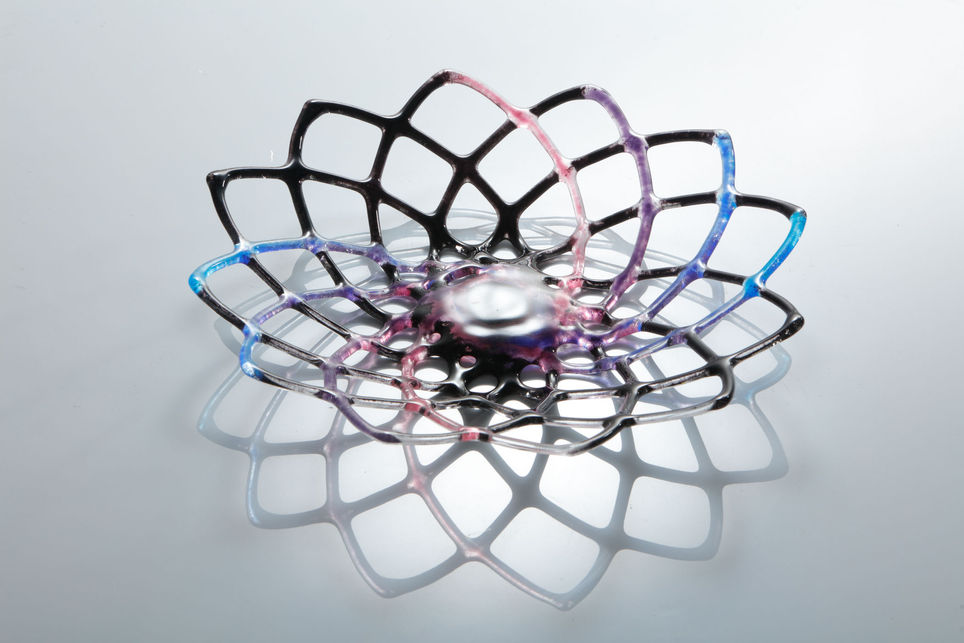 A glass hypotrochoid design sculpture on a table, glass is blue, pink, purple and black spiral galaxy, background is white.
