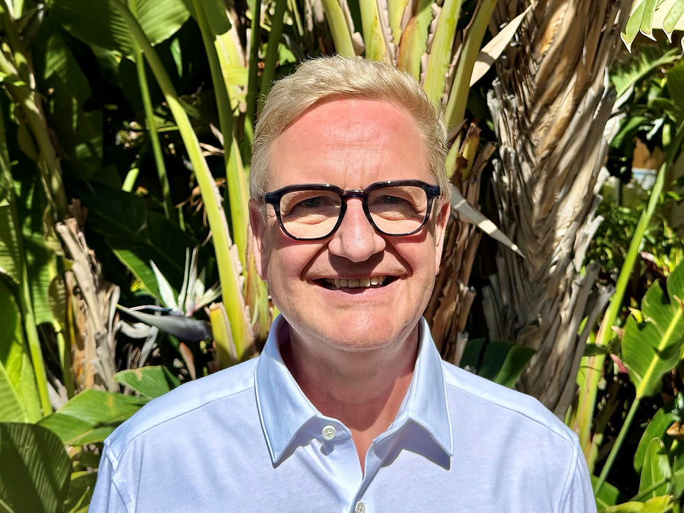 Smiling man in glasses and open-necked blue shirt with garden greenery behind.