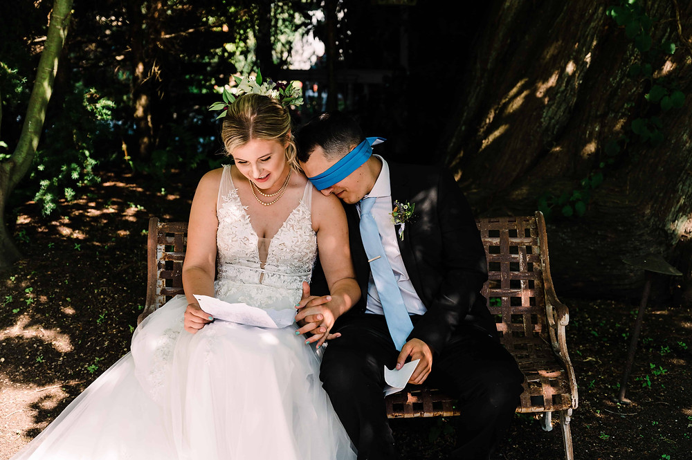 A blindfolded groom leans his forehead on his bride's shoulder as they sit next to each other, reading letters on their wedding day before the ceremony.