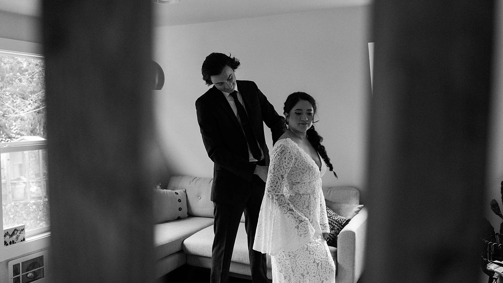 A tall man in a suit zips up the wedding dress of his bride as they get ready together for their elopement.