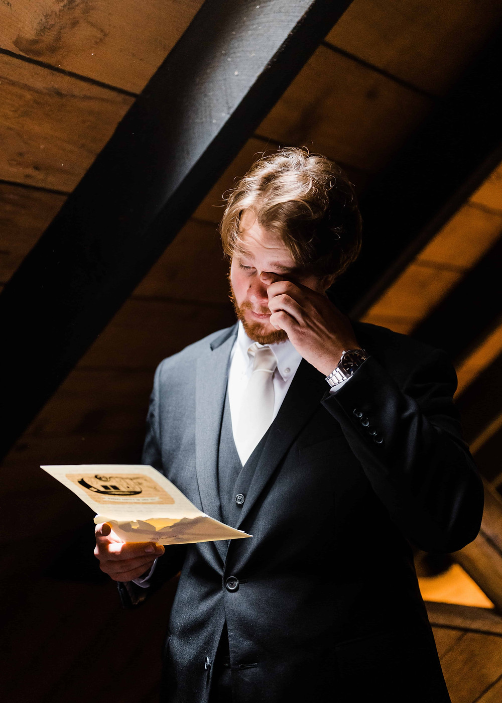 A man in a gray, three-piece suit wipes a tear from his eye as he reads a letter from his bride on their wedding day.