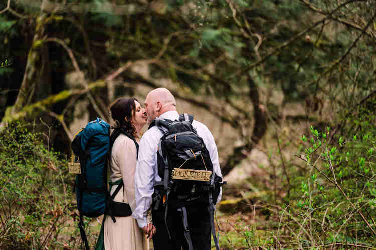 A bride and groom, wearing hiking backpacks and wedding clothes, lean in for a kiss surrounded by trees at their Royal Arch Park elopement