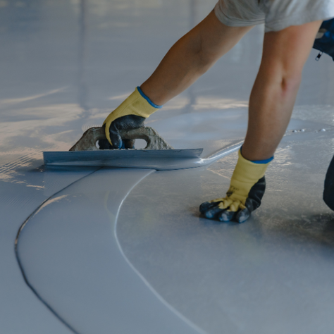 Worker kneels smoothing glossy gray floor coating with a trowel, wearing yellow gloves in a bright indoor space.