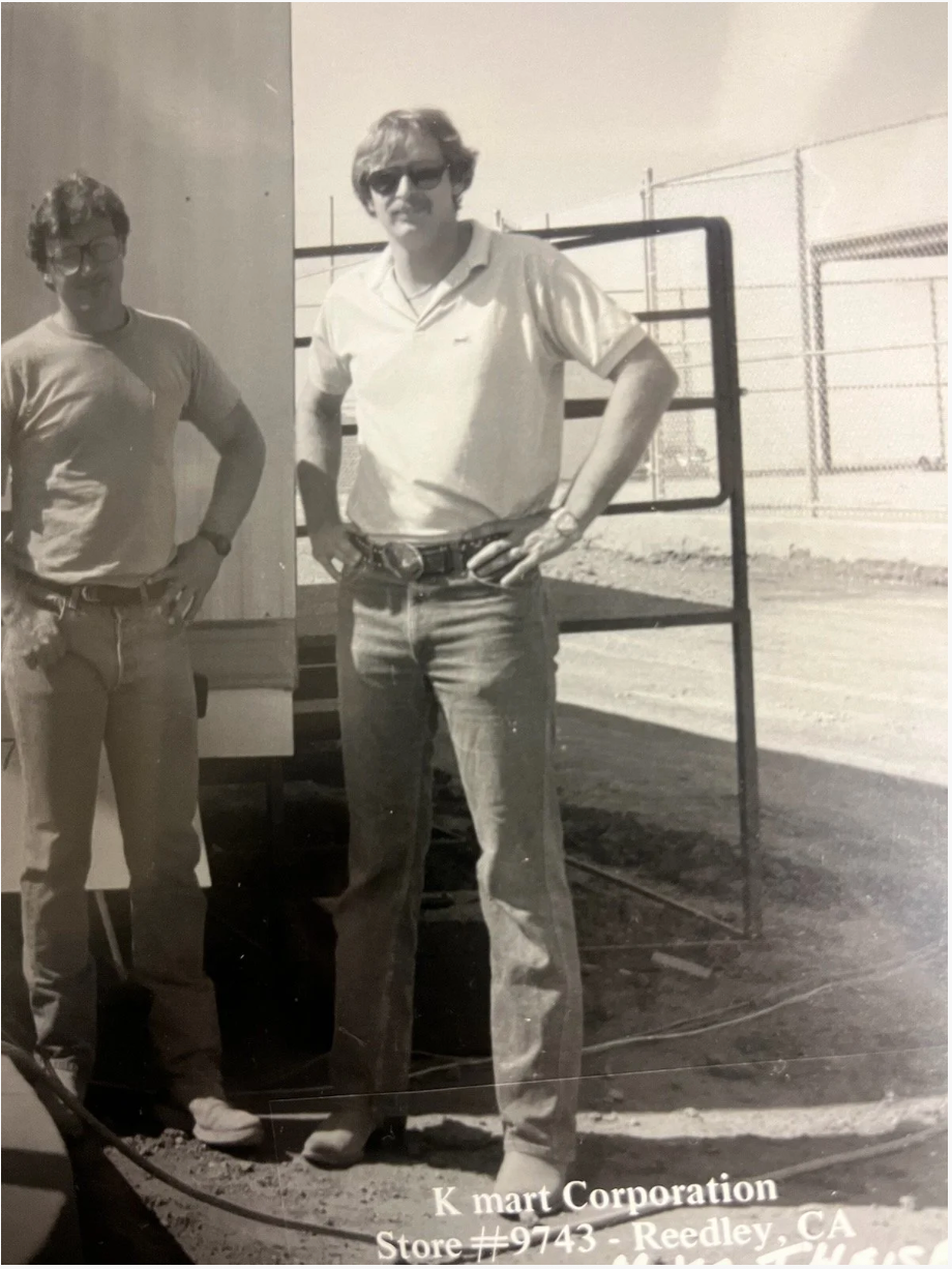 Two men stand confidently outdoors near a fence, wearing casual attire and sunglasses. Text: K mart Corporation, Store #9743, Reedley, CA.