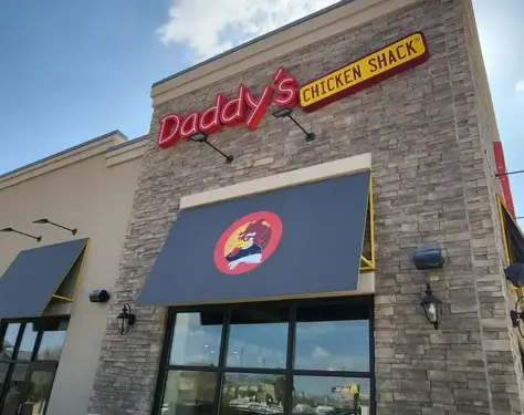 Exterior of Daddy’s Chicken Shack restaurant with stone facade, red neon sign, blue awnings, and clear sky.