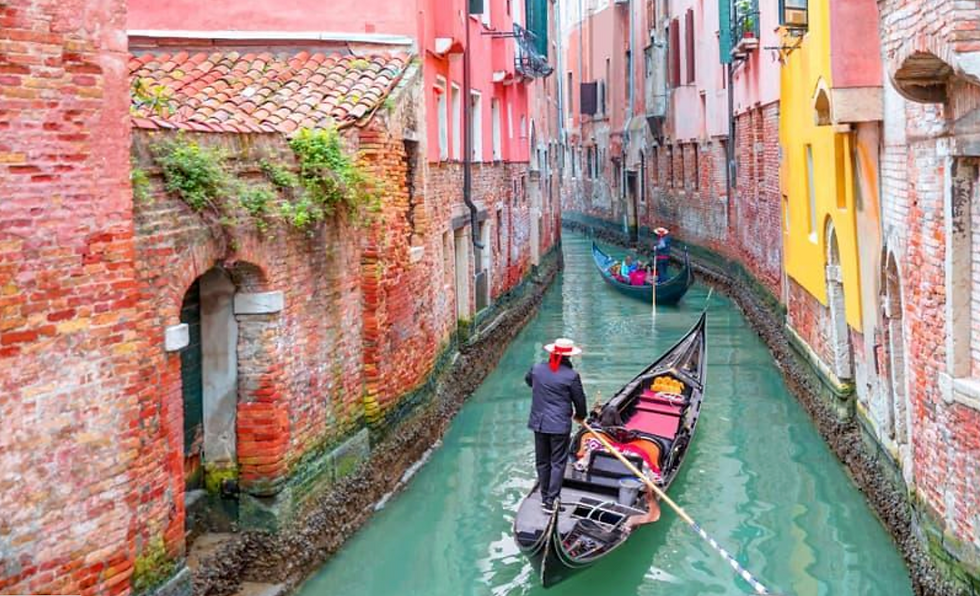 Gondolier steers a black gondola through a narrow Venetian canal beside colorful old buildings and brick walls.