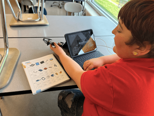 A student in a red shirt sits at a classroom table using a tablet to interact with a drone flight simulator. In front of them is a printed guide showing various drone components and controls. Sunlight streams through large windows in the background.