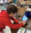 Young student wearing safety goggles interacts with a tablet while standing at a classroom table. A small training drone sits beside the tablet, and a colorful drone instruction booklet is open nearby—highlighting a hands-on STEM learning activity.