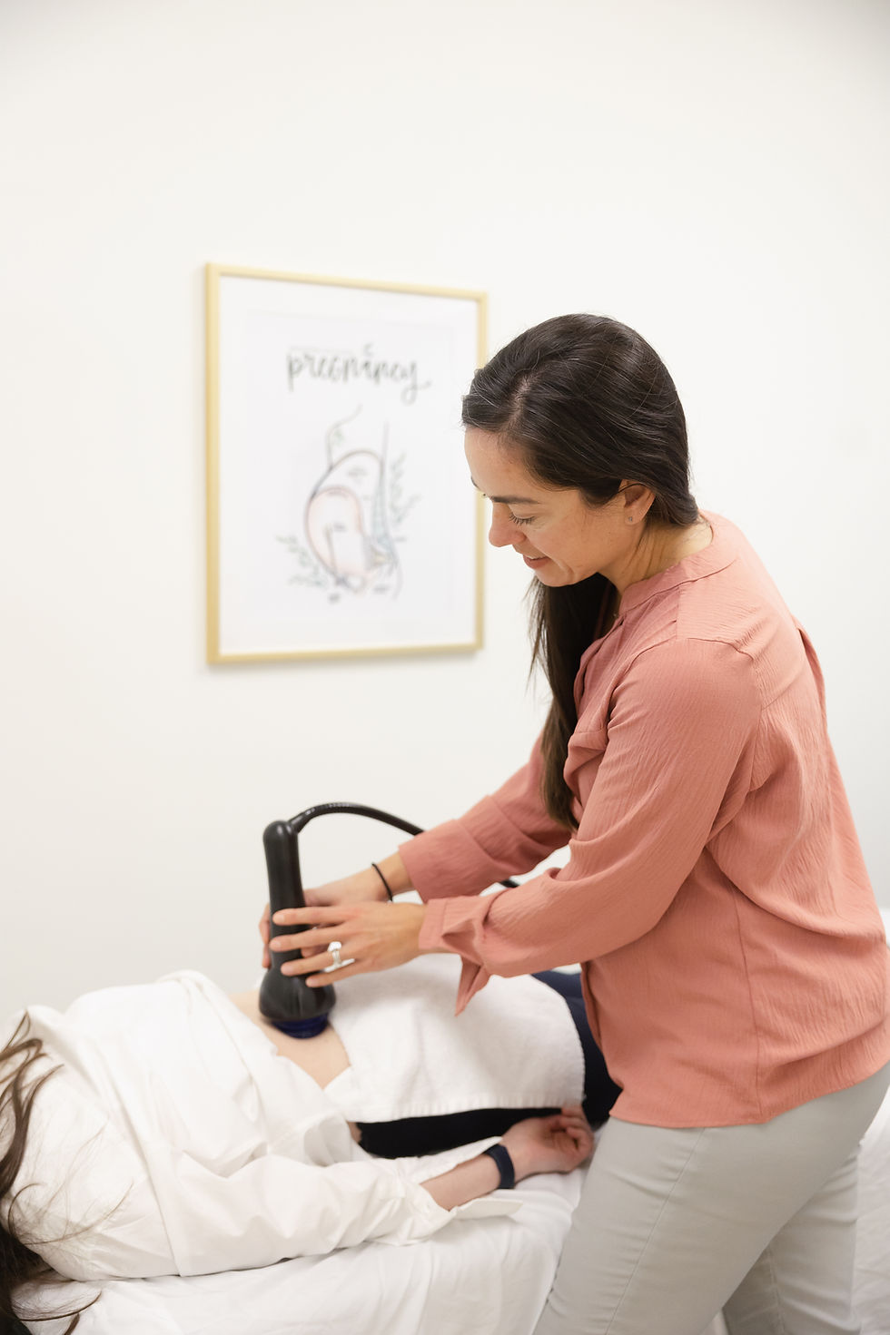 Woman in a pink blouse uses a handheld shockwave therapy device on a reclining patient in a bright clinic. Used for erectile dysfunction and pelvic pain.