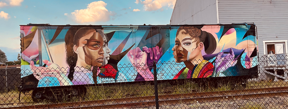 Colorful mural on a train car featuring portraits and abstract shapes. A dog peers out a window in the background. Fence in foreground.
