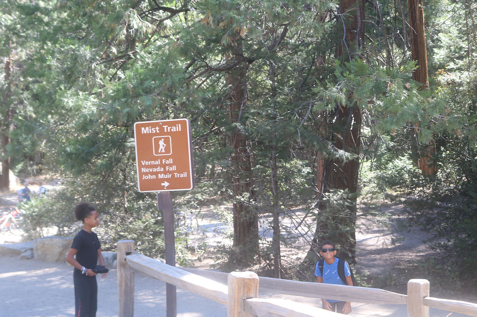 Two boys at a forest trail sign reading Mist Trail, Vernal Fall, Nevada Fall, John Muir Trail, with tall trees behind.
