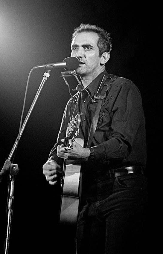 Paul Kelly standing at a microphone holding a guitar and a harmonica is around his neck