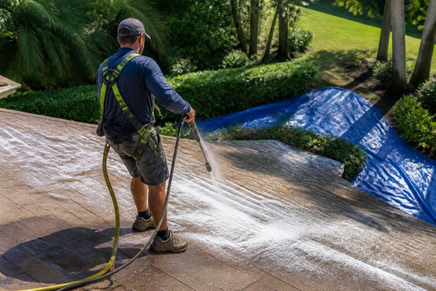 Technician using a soft wash system on shingles, affecting roof cleaning cost through protected landscaping.