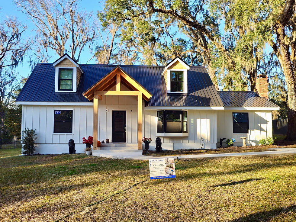 White house with black metal roof and wooden porch under a clear blue sky. Trees in the background. Sign on lawn reads DNA Painting.