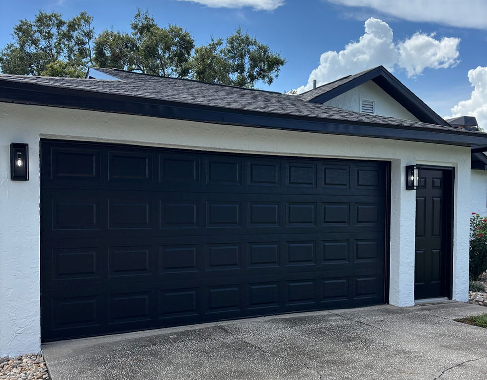 Black garage door and side door on a white house with black trims. Concrete driveway in front, trees, and a blue sky with clouds above.