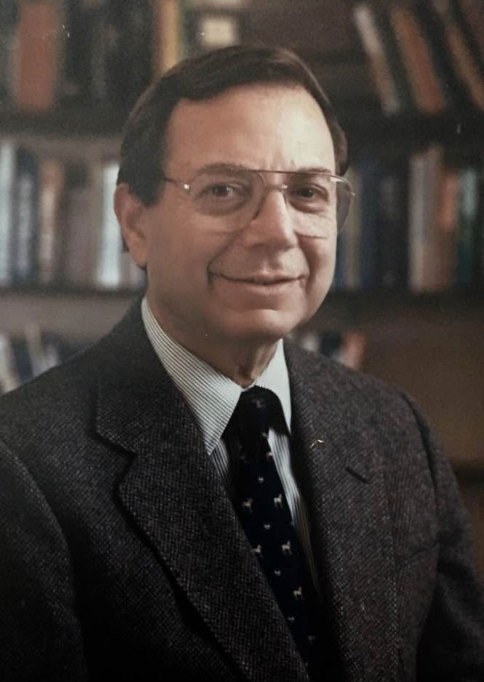 Smiling man in glasses and suit with bookshelves behind.