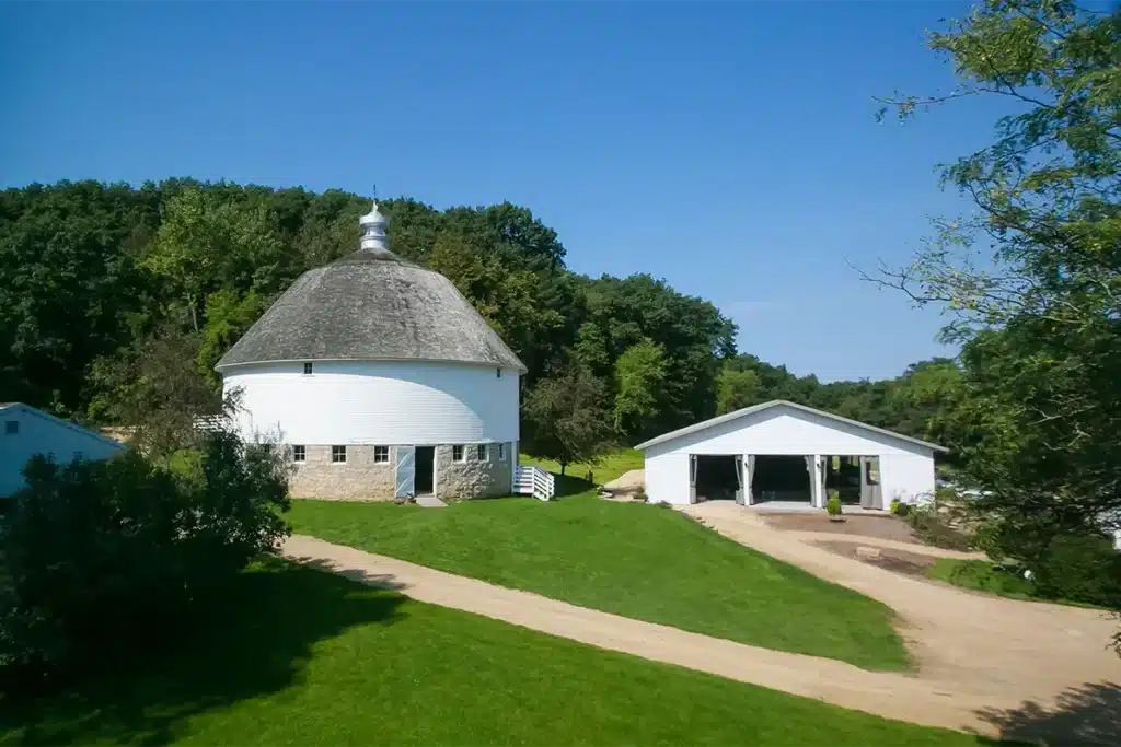 a white round barn nestled in the trees with green grass surrounding