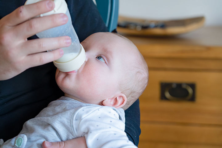 Baby being fed from a bottle by an adult, reclining in a chair indoors with a warm wooden background.
