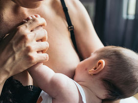 Mother breastfeeding baby in close-up, baby holding her chest in a warm, intimate moment by a window.