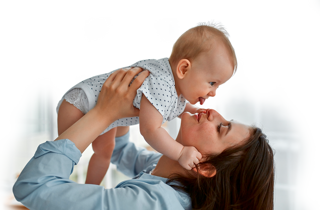 A woman is holding up her happy baby as they stick their tongue out